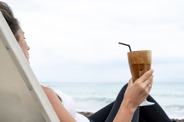 女士高加索女人拿着咖啡在沙滩上喝着泡沫饮料 拿着吸管喝着大海餐厅宁静沙滩