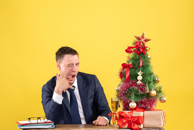 帅哥帅哥坐在圣诞树旁的桌子上对着相机眨眼睛 黄色的礼物坐着商人桌子