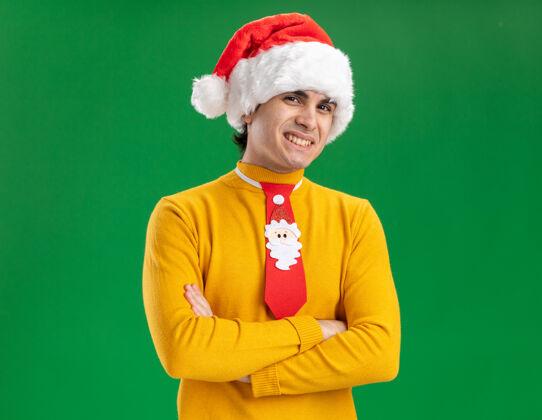 搞笑一个穿着黄色高领毛衣 戴着圣诞帽 打着滑稽领带的年轻人看着镜头 脸上挂着大大的笑容 双臂交叉着站在绿色的背景上看高领毛衣领带