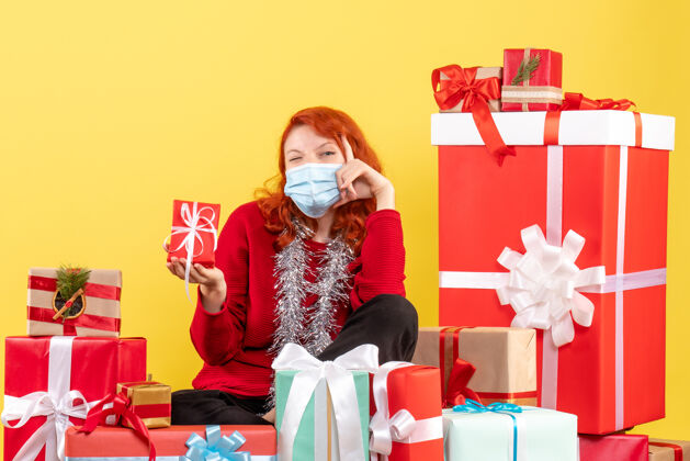 情感坐在黄色墙壁上戴着面具的年轻女子的圣诞礼物前视图购物周围礼物