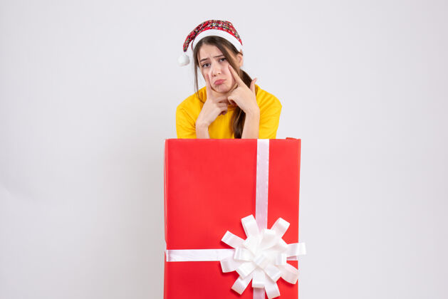 礼物戴着圣诞帽的悲伤女孩站在白色的大圣诞礼物后面悲伤的女孩圣诞老人站着