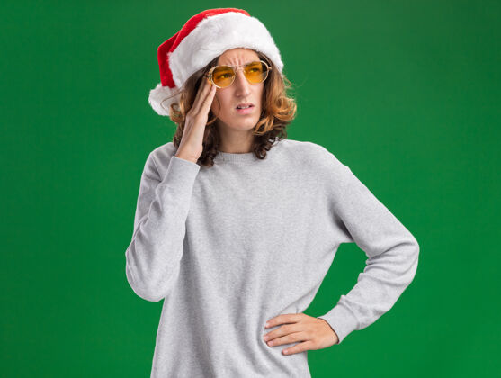 圣诞老人戴着圣诞老人帽 戴着黄色眼镜的年轻人站在绿色的背景下 茫然地看着一边年轻人一旁站着