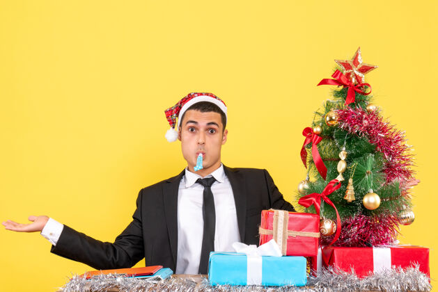 男人正面图：戴着圣诞帽的年轻人坐在桌子旁 张开双手 用噪音制造圣诞树和礼物公司年轻人圣诞节