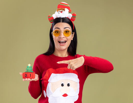 橄榄印象深刻的年轻白人女孩戴着圣诞老人的头带和毛衣与眼镜举行和指着圣诞树玩具与日期隔离在橄榄绿的墙上圣诞节圣诞老人日期