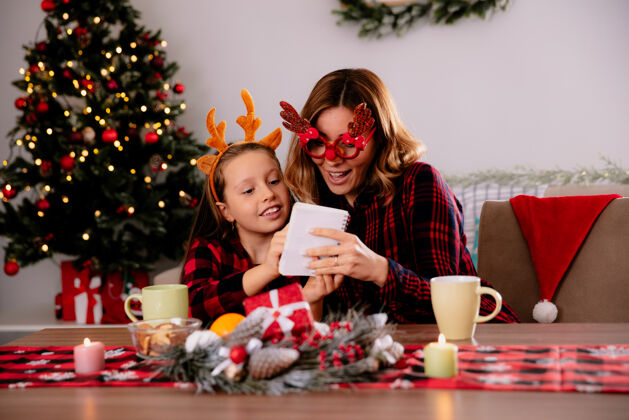圣诞节妈妈戴着驯鹿眼镜看着笔记本 女儿坐在桌旁 在家里享受圣诞节的时光印象深刻享受女儿
