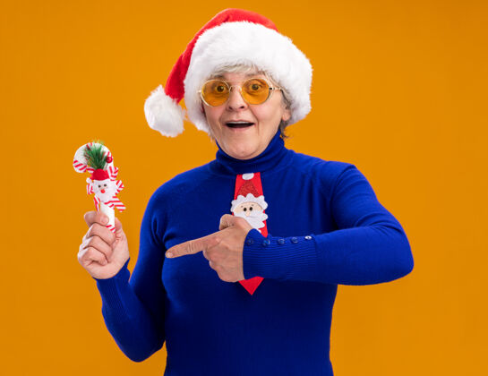 印象深刻一位戴着太阳眼镜 戴着圣诞帽 系着圣诞领带的老妇人站着 指着橙色背景上的糖果手杖 手杖上留有复印空间领带老人手杖