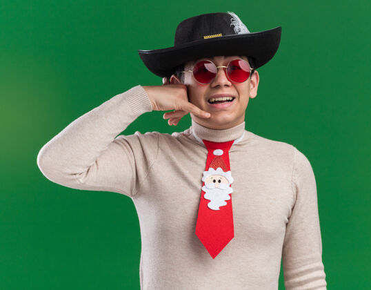 领带微笑的年轻人戴着帽子 打着圣诞领带 戴着眼镜 在绿色的墙上显示着打电话的手势穿着展示电话