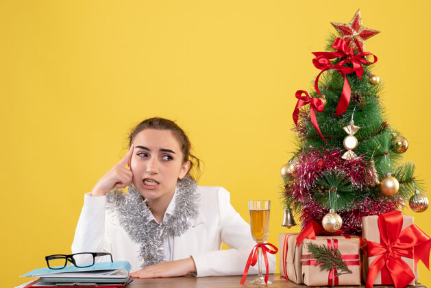 女医生正面图：女医生坐在桌子后面 黄色背景上有圣诞树和礼品盒微笑背景坐着