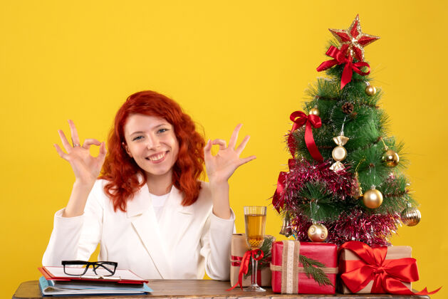 圣诞节正面图：女医生坐在桌子后面 拿着圣诞礼物 黄色背景上的树女医生插花健康