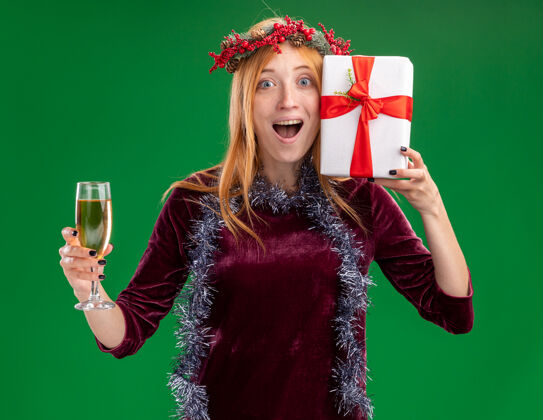 圣诞节快乐的年轻漂亮的女孩穿着红色的裙子 脖子上戴着花环和花环 手里拿着一杯香槟酒 绿色的墙上隔着一个礼盒香槟玻璃杯花环