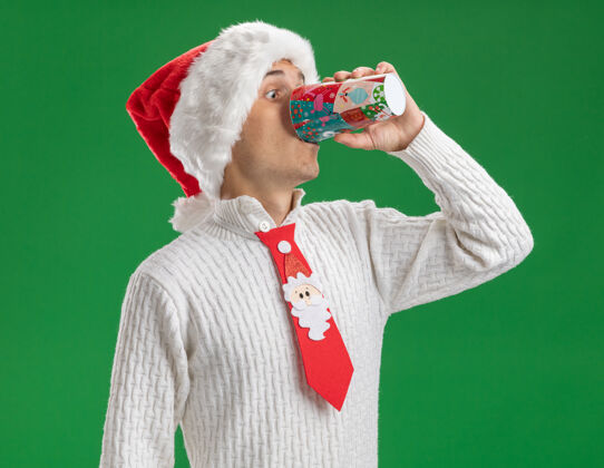 咖啡给人印象深刻的年轻帅哥戴着圣诞帽打着圣诞老人的领带喝着咖啡从塑料圣诞杯隔离在绿色的墙上年轻圣诞老人喝酒