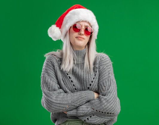 眼镜愤愤不平的年轻金发女人 穿着冬季毛衣 戴着圣诞老人帽 戴着红眼镜 皱着眉头 双臂交叉站在绿色背景上 看着镜头帽子脸皱眉