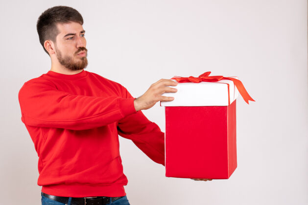 年轻男性身穿红衫的年轻人在白色墙上的盒子里拿着圣诞礼物的正视图人节日快乐