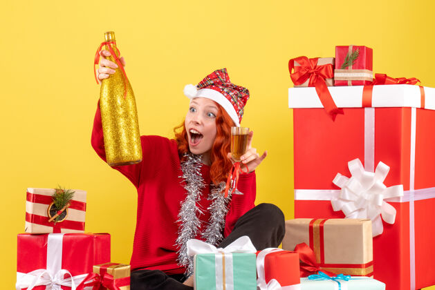 人年轻女子坐在圣诞礼物周围 在黄色的墙上用香槟庆祝的正面视图庆祝购物前面