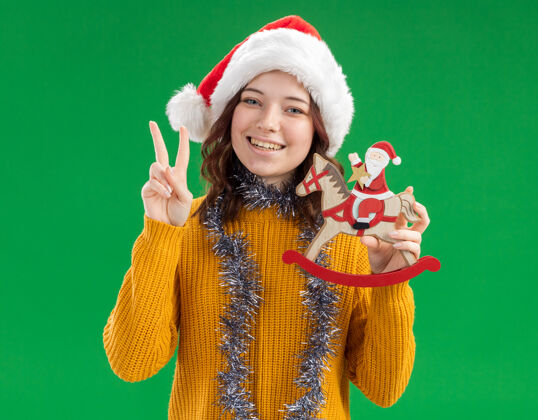 斯拉夫微笑的年轻斯拉夫女孩 戴着圣诞帽 脖子上戴着花环 把圣诞老人抱在摇马装饰上 并做着胜利的手势脖子圣诞老人花环