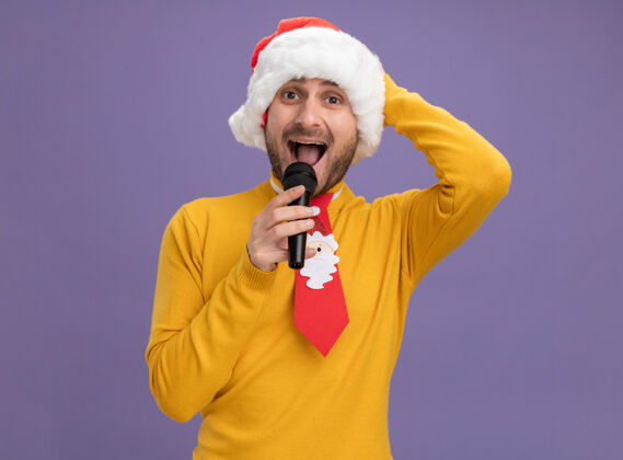 年轻令人印象深刻的年轻白人男子戴着圣诞帽 打着领带 手持麦克风近嘴 手放在头上 隔离在紫色的墙上靠近保持帽子