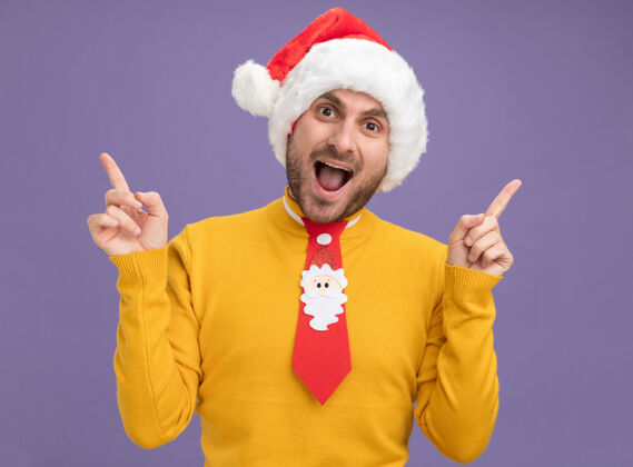 圣诞节令人印象深刻的年轻白人男子戴着圣诞帽 打着领带指着隔离在紫色墙上指着男人帽子