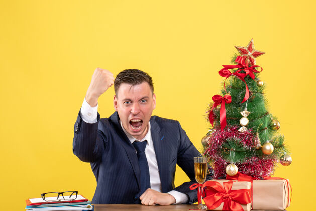商人前视图兴高采烈的男子显示获胜的姿态坐在圣诞树附近的桌子上 黄色的礼物办公室黄色手势