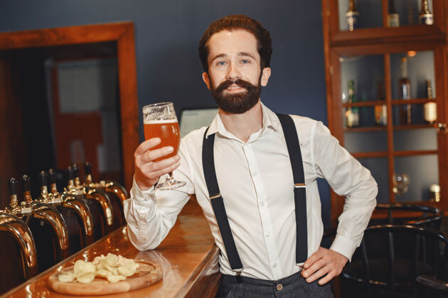 领带一个留着胡子的男人站在吧台旁 喝着酒杯里的酒啤酒厂酒吧成人
