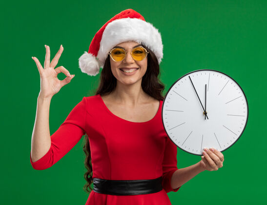 漂亮快乐的年轻漂亮女孩戴着圣诞帽 戴着眼镜 拿着时钟 看着相机 在绿色背景上做着“ok”标志抱着时钟圣诞老人