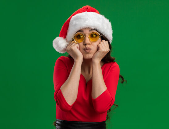 无聊无聊的年轻漂亮女孩戴着圣诞帽 戴着眼镜 手放在下巴上鼓起的脸颊上 看着绿色背景上的隔离边 还有复制空间下巴年轻漂亮