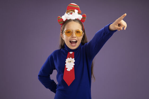 高领毛衣小女孩穿着蓝色高领毛衣 系着红色领带 头上戴着有趣的圣诞戒指 高兴而兴奋地看着相机 食指指向紫色背景上的一边女孩搞笑手指