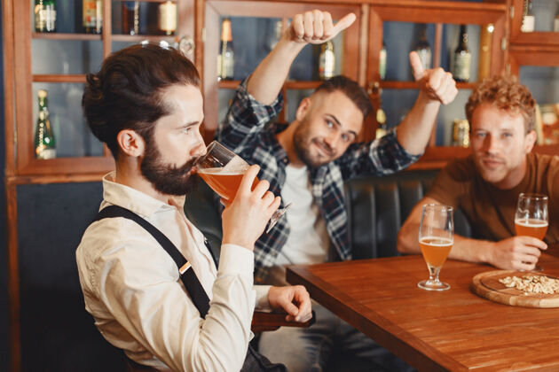 啤酒和最好的朋友见面三个穿着休闲服的快乐的年轻人一起坐在酒吧里聊天喝啤酒年轻酒吧酒吧
