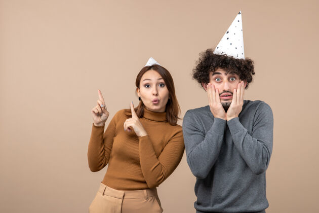 年轻可爱兴奋的年轻夫妇戴着新年帽的女孩指着上方 站在灰色图片上的男人身边站着帽子大人