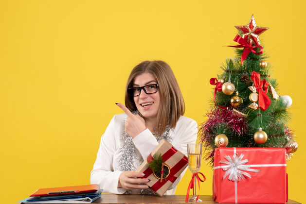 办公室正面图：女医生坐在桌子前 手里拿着黄色背景的礼物 还有圣诞树和礼品盒微笑健康导演