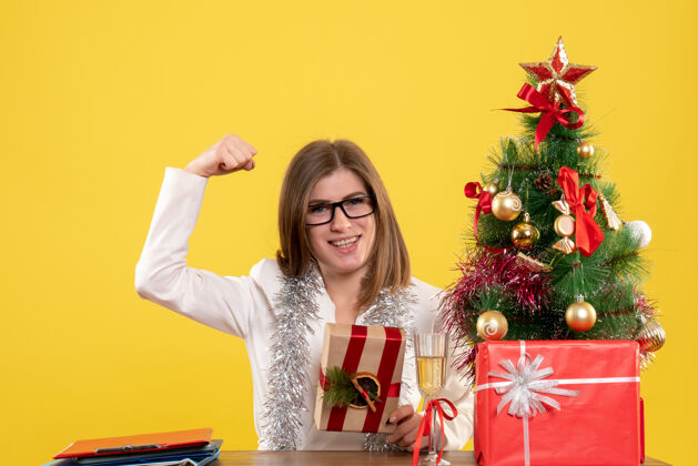 现在正面图：女医生坐在桌子前 手里拿着黄色背景的礼物 还有圣诞树和礼品盒桌子背景前面