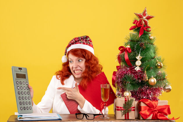 庆祝正面图：女医生坐在桌子后面 拿着黄色背景的计算器 还有圣诞树和礼品盒餐桌圣诞节医生