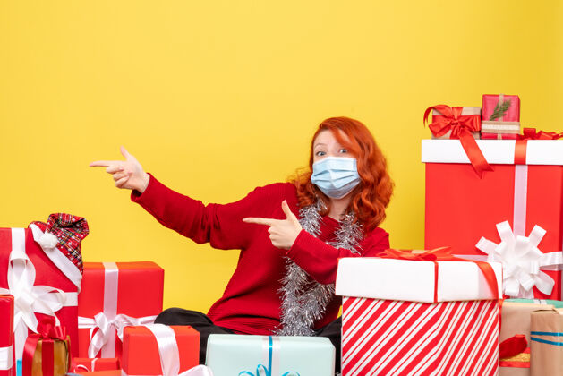 坐着一个戴着面具坐在黄色墙壁上的年轻女子围着圣诞礼物的正视图病毒面具女士