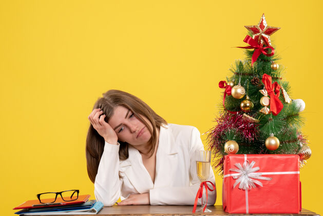 圣诞节正面图：女医生坐在黄色背景的桌子前 放着圣诞树和礼品盒坐人医生