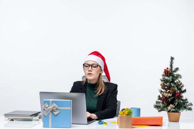 圣诞树戴着圣诞老人帽的女商人坐在桌子旁 桌上放着圣诞树和礼物电脑女人圣诞老人