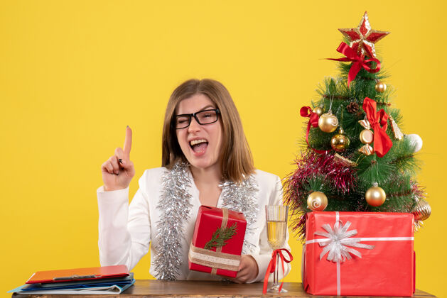 坐正面图：女医生坐在桌子前 手里拿着黄色背景的礼物 还有圣诞树和礼品盒办公室举行医生
