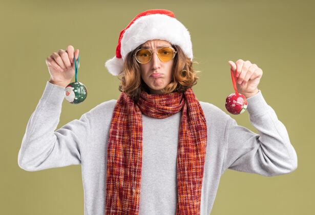 相机沮丧的年轻人戴着圣诞老人的帽子 戴着黄色的眼镜 脖子上围着暖和的围巾 手里拿着圣诞球 看着相机 脸上带着悲伤的表情 噘着嘴唇站在绿色的背景上抱着站着表情