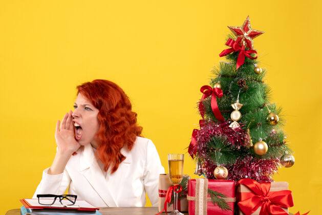 女医生前视图：女医生坐在桌子后面 手里拿着圣诞礼物 黄色背景上的圣诞树在呼唤前面健康快乐