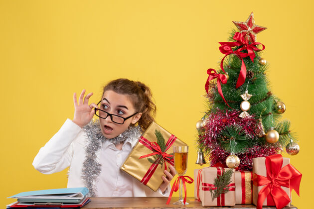 人前视图：女医生坐在桌子后面 拿着圣诞礼物 黄色背景上的树圣诞节前面女性