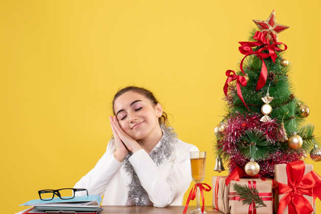 圣诞节正面图：女医生坐在桌子后面 脸上露出喜悦的表情 背景是黄色的圣诞树和礼盒桌子情绪背景