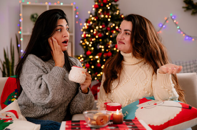 困惑焦急的年轻女孩拿着杯子 看着困惑的朋友坐在扶手椅上 在家里享受圣诞节时光女孩看美丽