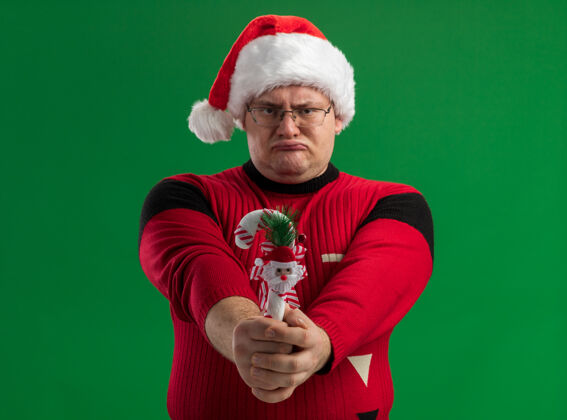 手杖愁眉不展的成年男子戴着眼镜和圣诞帽 向镜头伸出糖果手杖装饰物 看着隔离在绿色背景上的镜头圣诞快乐帽子皱眉