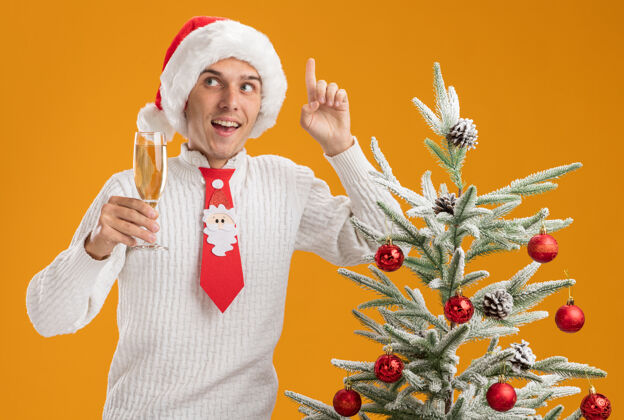 年轻印象深刻的年轻帅哥戴着圣诞帽 打着圣诞老人的领带 站在装饰好的圣诞树旁 手里拿着一杯香槟 看着一边指着橙色的墙上孤立的杯子印象深刻指着