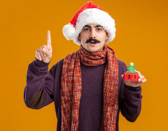 温暖留着胡子的男人戴着圣诞老人帽 脖子上围着暖和的围巾 手里拿着新年礼物的玩具立方体 自信地用食指站在橙色的背景上站立新的索引