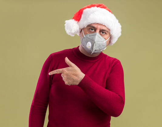 保护中年男子戴着圣诞老人帽戴着护面面具看着相机严肃的脸用食指指着旁边站在绿色背景上严重帽子食指