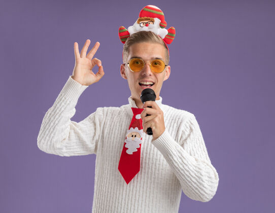 麦克风印象深刻的年轻帅哥戴着圣诞老人的头带和领带戴着眼镜拿着麦克风近嘴做ok标志隔离在紫色的墙上圣诞老人年轻头带