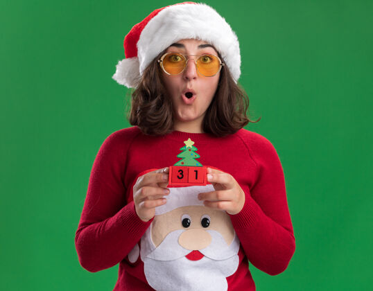 圣诞老人穿着圣诞毛衣 戴着圣诞帽 戴着圣诞眼镜 手里拿着新年礼物的玩具立方体的小女孩站在绿色的墙边 惊讶不已毛衣圣诞节立方体