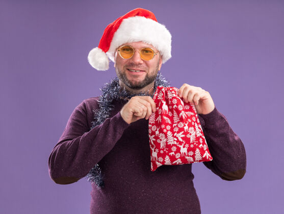 圣诞节高兴的中年男子戴着圣诞帽 脖子上戴着金箔花环 戴着眼镜 手里拿着圣诞礼品袋 隔离在紫色的墙上脖子帽子戴着