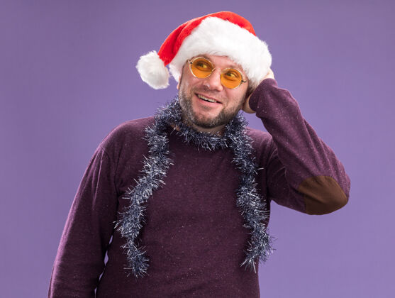 感动快乐的中年男子戴着圣诞帽 脖子上戴着金箔花环 戴着眼镜 看着隔离在紫色墙上的头中年帽子脖子
