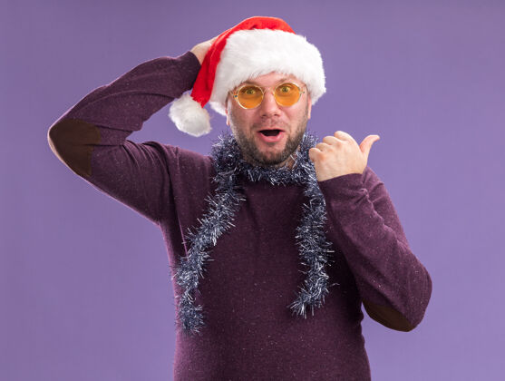 男人令人印象深刻的中年男子戴着圣诞帽 脖子上戴着金箔花环 戴着眼镜 手放在头上 指着紫色墙壁上孤立的一面帽子周围圣诞老人