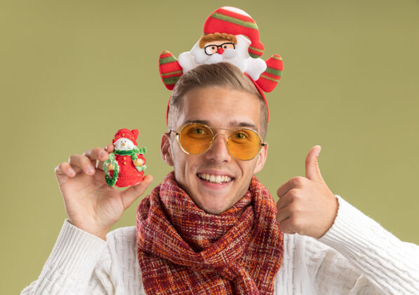 拇指快乐的年轻帅哥戴着圣诞老人的头带和围巾 手里拿着雪人的圣诞饰品 竖起大拇指 孤零零地站在橄榄绿的墙上穿着雪人年轻
